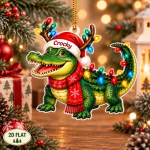 Könnte beinhalten: Ein flacher, hängender Weihnachtsbaumschmuck in Form eines grünen Krokodils, das eine rote Weihnachtsmütze mit dem Namen Crocky auf dem Rand und einen roten Schal mit weißen Schneeflocken trägt. Das Krokodil ist mit bunten Lichterketten verziert.