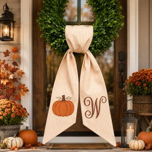 May include: A green boxwood wreath adorned with a long, beige and white striped fabric sash. The sash features an embroidered orange pumpkin on the left tail and a large, brown, cursive letter W on the right tail, hanging over a dark wooden door.