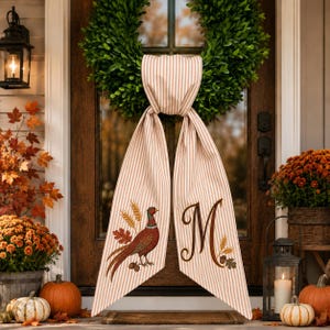 May include: A green boxwood wreath hanging on a door, adorned with a long, beige and white striped fabric sash. The sash features a printed illustration of a pheasant with wheat stalks on one side and a large, brown, cursive letter M on the other.