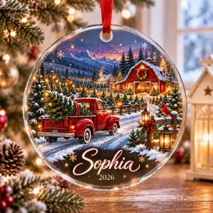May include: A round, clear glass Christmas ornament featuring a printed winter scene with a red pickup truck carrying a pine tree, a red barn, snow-covered mountains, and a cardinal. The name Sophia and the year 2026 are printed in white script.
