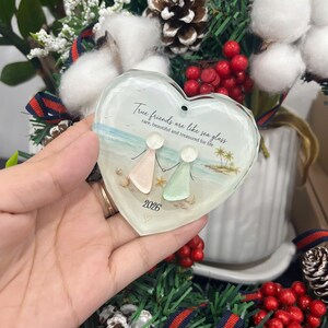 May include: A heart-shaped glass ornament featuring a beach scene with two figures made of sea glass, a starfish, and palm trees. The text reads: True friends are like sea glass, rare, beautiful and treasured for life, with the year 2026 below.