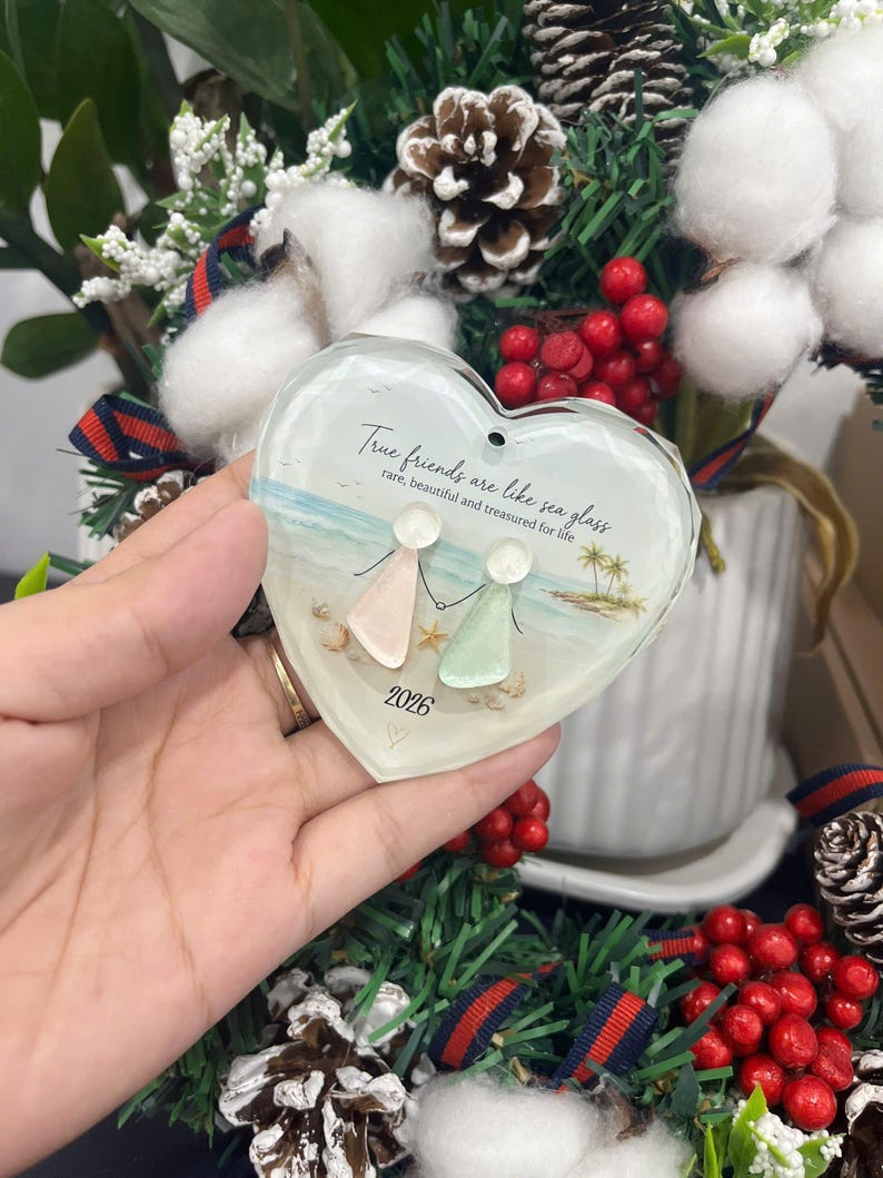 May include: A heart-shaped glass ornament featuring a beach scene with two figures made of sea glass, a starfish, and palm trees. The text reads: True friends are like sea glass, rare, beautiful and treasured for life, with the year 2026 below.