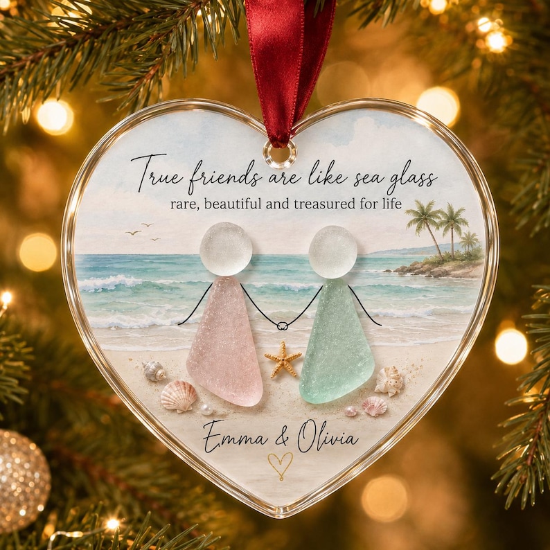 May include: A heart-shaped glass ornament featuring a beach scene with two figures made of pink and light green sea glass holding hands. The text reads True friends are like sea glass, rare, beautiful and treasured for life, Emma & Olivia.