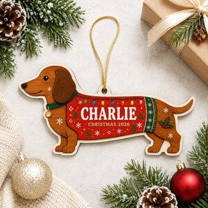 May include: A wooden dachshund ornament featuring a red sweater with green trim, decorated with colorful Christmas lights, snowflakes, and holly. The sweater displays the text CHARLIE and CHRISTMAS 2026 in white. A gold cord is attached for hanging.