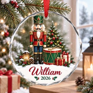 Può includere: Un addobbo natalizio rotondo in vetro trasparente con uno schiaccianoci in uniforme rossa e verde, un albero di Natale decorato, un tamburo e regali. Il testo William e 2026 è stampato in rosso e verde nella parte inferiore.