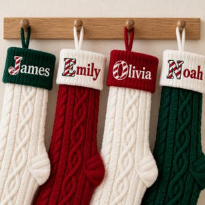 May include: Four cable-knit Christmas stockings hanging from a wooden rack. The stockings are in alternating red, white, and dark green colors with contrasting cuffs. Each cuff features a personalized name: James, Emily, Olivia, and Noah.
