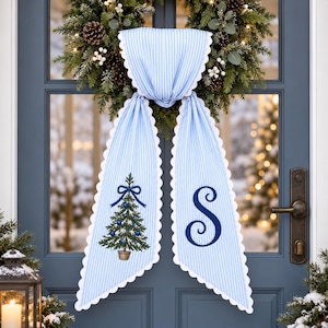 Peut inclure: Une suspension de porte en tissu à rayures bleu clair et blanc avec des bords festonnés. La partie gauche présente un sapin de Noël brodé avec des ornements bleus et un nœud, tandis que la partie droite affiche une grande lettre S brodée.