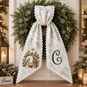 May include: A decorative fabric swag featuring a light green and white floral pattern with scalloped edges. One side displays a printed nativity scene with a star, while the other side features a large, dark green embroidered letter C.