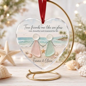 May include: A heart-shaped clear acrylic ornament hanging from a gold metal stand by a red ribbon. The ornament features a beach scene with two figures made of sea glass, one pink and one green, holding hands. Text reads: True friends are like sea glass, rare, beautiful and treasured for life, Emma & Olivia.