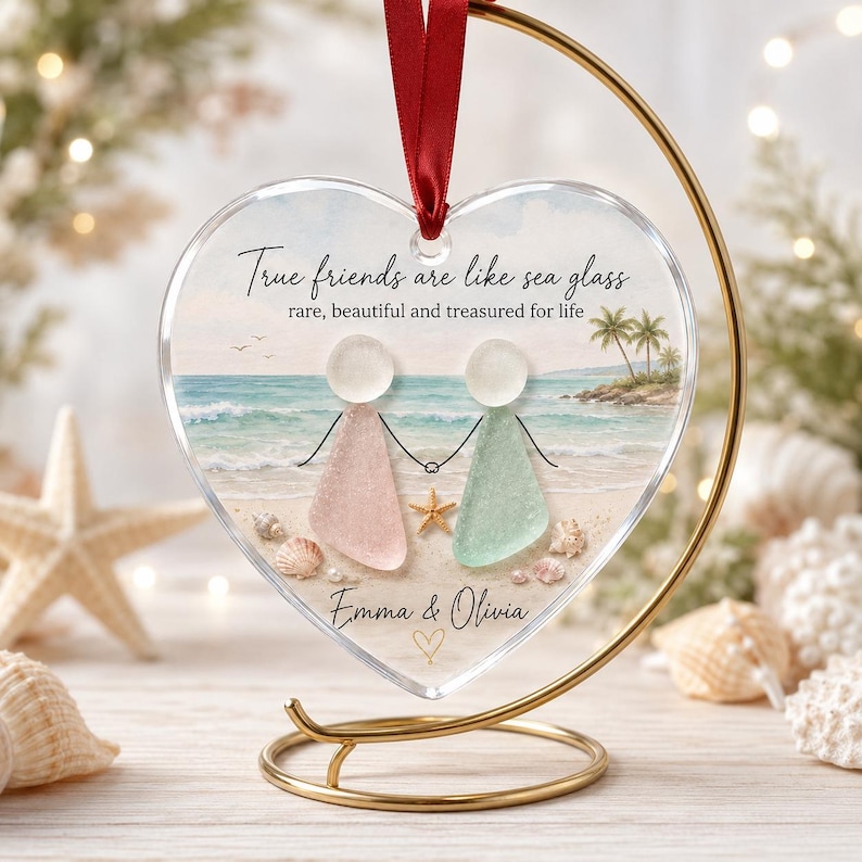 May include: A heart-shaped clear acrylic ornament hanging from a gold metal stand by a red ribbon. The ornament features a beach scene with two figures made of sea glass, one pink and one green, holding hands. Text reads: True friends are like sea glass, rare, beautiful and treasured for life, Emma & Olivia.
