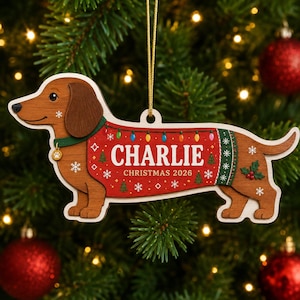 May include: A wooden dachshund ornament featuring a brown dog wearing a red and green sweater with the text CHARLIE and CHRISTMAS 2026. The sweater includes a string of colorful lights, snowflakes, and small trees. A gold string is attached for hanging.