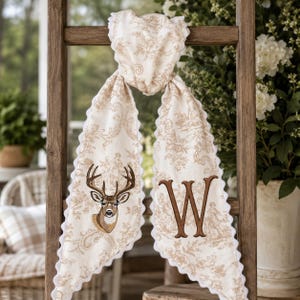 May include: A cream-colored fabric scarf featuring a tan toile pattern, a scalloped white edge, an embroidered brown deer head on the left tail, and a large embroidered brown letter W on the right tail, draped over a wooden ladder rung.