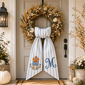 May include: A round autumn wreath featuring dried wheat, grasses, and small white pumpkins. A large, light blue and white striped fabric bow hangs from the center, with a stack of three pumpkins on one tail and a blue embroidered letter M on the other.