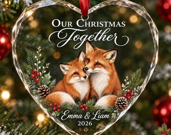 Può includere: Una decorazione natalizia in acrilico trasparente a forma di cuore con un'illustrazione stampata di due volpi accoccolate, circondate da rami di pino, bacche rosse e pigne. Il testo recita Our Christmas Together, Emma & Liam, 2026.