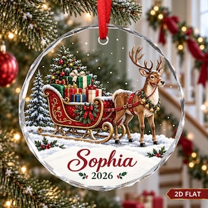 May include: A circular glass Christmas ornament featuring a red sleigh filled with wrapped gifts, pulled by a reindeer wearing a bell collar. The design includes snow, pine trees, and the text "Sophia 2026" in red and green script.
