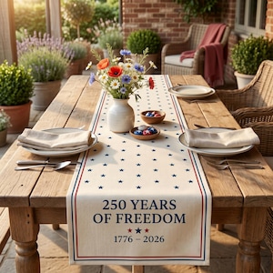 May include: A wooden table set for a meal, featuring a cream-colored table runner with blue stars and the text "250 YEARS OF FREEDOM 1776-2026." The table is set with plates, silverware, and a vase of flowers.