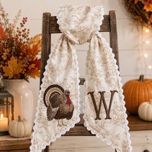 Pu&ograve; includere: Un runner in tessuto color crema con motivo toile beige, bordo smerlato bianco e un tacchino ricamato in marrone su un lato. L'altro lato presenta una grande lettera W ricamata in marrone con carattere serif, centrata sul tessuto.