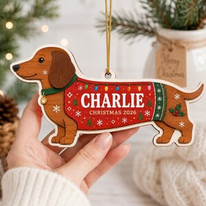May include: A wooden dachshund ornament featuring a brown dog wearing a red sweater with green trim. The sweater displays the text CHARLIE and CHRISTMAS 2026, decorated with colorful light bulbs, snowflakes, and small trees. A gold string is attached.
