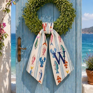 May include: A green wreath hangs on a light blue door with a brass handle. A decorative scarf with a fish and lobster pattern is tied to the wreath. The scarf has blue stripes and a large navy blue "W" on it. The background shows the ocean.