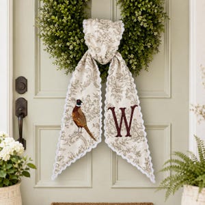 May include: A decorative fabric door hanger featuring a beige toile pattern with a pheasant illustration on the left side and a large, dark red embroidered letter W on the right side, finished with a white scalloped trim edge.