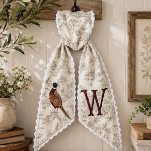 May include: A hanging fabric towel featuring a cream-coloured base with a grey floral toile pattern and white scalloped trim. One side displays a detailed illustration of a pheasant, while the other side features a large, embroidered letter W in dark red.