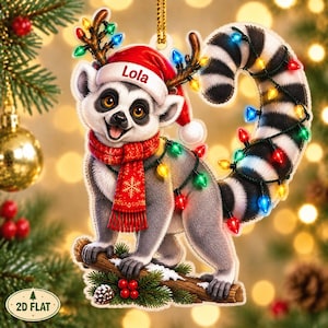 Könnte beinhalten: Ein flacher Weihnachtsbaumanhänger mit einem Lemuren, der eine rote Weihnachtsmütze mit dem Namen Lola, einen roten Schal und ein mit bunten Lichterketten geschmücktes Geweih trägt. Der Lemur hat einen langen, geringelten Schwanz mit Lichtern.
