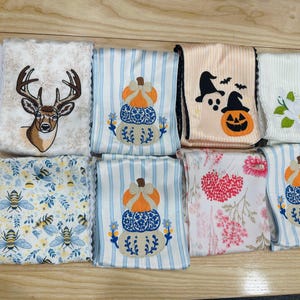May include: A collection of eight folded fabric towels featuring various embroidered designs including a deer head, stacked pumpkins, ghosts with a jack-o'-lantern, a white flower, bees with blue leaves, and red floral patterns on white and striped backgrounds.
