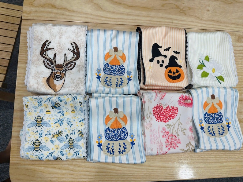 May include: A collection of eight folded fabric towels featuring various embroidered designs including a deer head, stacked pumpkins, ghosts with a jack-o'-lantern, a white flower, bees with blue leaves, and red floral patterns on white and striped backgrounds.