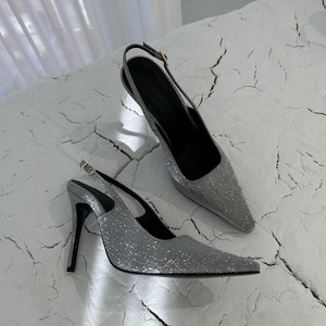 以下が含まれることがあります： ポインテッドトゥとハイヒールが特徴の、シルバーグリッターのスリングバックヒール。靴はバックル留めと黒いソールが付いています。ヒールは、質感のある白い表面に置かれています。