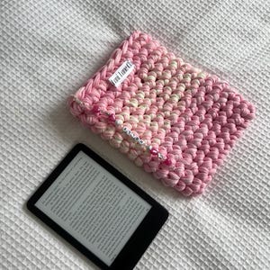 May include: A rectangular, hand-crocheted sleeve in shades of pink and white, featuring a white fabric tag labelled "Lani Lane Co." and a decorative beaded charm strand with the name "HOLLY" in white letter beads, next to a black e-reader.