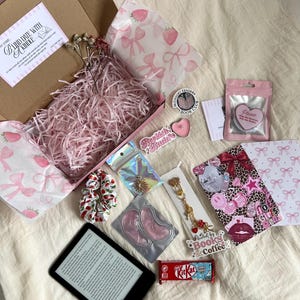 May include: A gift box filled with pink paper shred, a strawberry-patterned tissue paper, a black e-reader, a KitKat bar, a gold-toned charm keychain, pink under-eye patches, a cherry-print scrunchie, stickers, and a pink heart-shaped tin.
