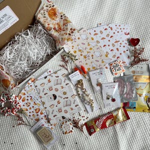 May include: A gift box containing autumn-themed items including patterned paper with pumpkins and geese, a pearl and gold-tone charm, a coffee cup charm, a hair scrunchie, a hydrogel face mask, a pack of Maltesers, and various small decorative stickers.