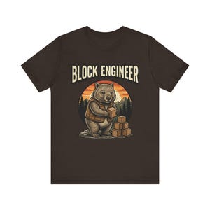 Könnte beinhalten: Dunkelbraunes T-Shirt mit einem Cartoon-Wombat, der eine Weste trägt und Holzblöcke hält. Der Wombat ist vor einem Sonnenuntergang und einer Bergkulisse platziert. Der Text "BLOCK ENGINEER" ist über dem Wombat gewölbt.