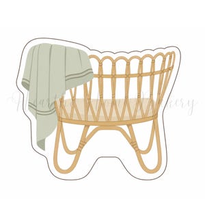 May include: A light brown, wicker-style baby cradle with a curved base and a soft green blanket draped over the side. The cradle has a woven design and is set against a white background.