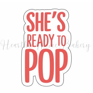 Puede incluir: Un cortador de galletas rojo y blanco con el texto "SHE'S READY TO POP" en una fuente en negrita y redondeada. El texto está en forma rectangular con esquinas redondeadas.