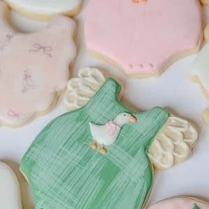 May include: A selection of decorated sugar biscuits in the shape of baby clothes. The biscuits are iced in pastel colours, including pink, green, and white. Each biscuit is decorated with a different design, such as a goose or a bow.