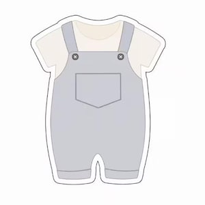 Può includere: Una tutina stile salopette grigio chiaro con sotto una maglietta a maniche corte color crema. Il capo presenta due bottoni grigio scuro sulle bretelle anteriori, una tasca centrale sul petto e un bordo bianco che ne delinea la forma.