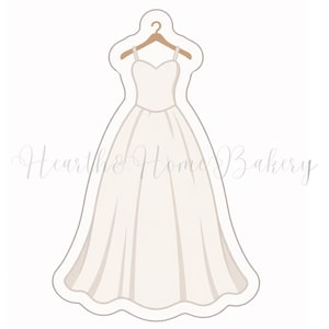 Peut inclure: Emporte-pièce en forme de robe de mariée blanche avec un décolleté en cœur et des bretelles spaghetti, suspendue à un cintre en bois. La robe a une jupe ample et est soulignée en beige clair. Le texte "Heart & Home Bakery" est écrit en cursive.