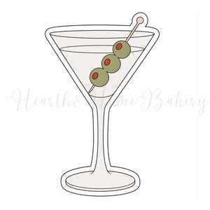 May include: A martini glass cookie cutter with a stem and base, filled with a light-colored liquid and garnished with three olives on a toothpick. The olives are green with red centers. The cookie cutter is white.