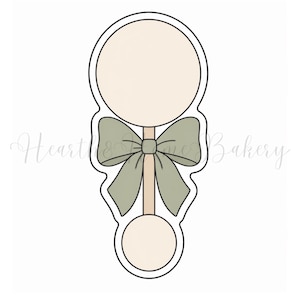 May include: A cookie cutter in the shape of a baby rattle. The rattle has a large circle at the top and a smaller circle at the bottom, connected by a stick. A sage green bow is tied around the stick. The rattle is beige with a black outline.