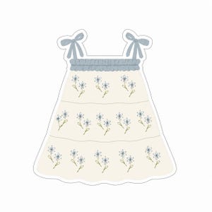 Puede incluir: Ilustración de un vestido de color claro con bordado floral azul. El vestido presenta un escote con volantes, tirantes con lazo y una falda escalonada. El diseño está delineado con un borde blanco.