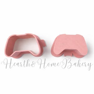 Puede incluir: Dos cortadores de galletas rosas con forma de mandos de videojuegos. Uno es un contorno, el otro una forma sólida. El texto en cursiva "Hearth & Home Bakery" está escrito debajo.