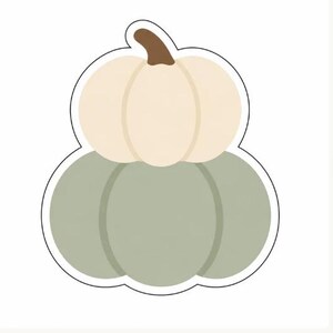 Può includere: Un adesivo con un design a due zucche sovrapposte. La zucca superiore è color crema con un picciolo marrone, mentre quella inferiore è verde salvia. L'intera illustrazione presenta un bordo bianco spesso che ne delinea la forma.