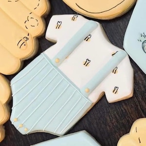 May include: A baby onesie cookie decorated with light blue suspenders and accents. The white onesie has small bee designs. Other cookies in the image include a yellow cloud and a light blue cookie with a design.