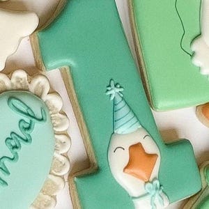 May include: A teal-colored cookie shaped like the number one, decorated with a cartoon goose wearing a party hat and bow. The cookie has a light brown border and is surrounded by other decorated cookies.