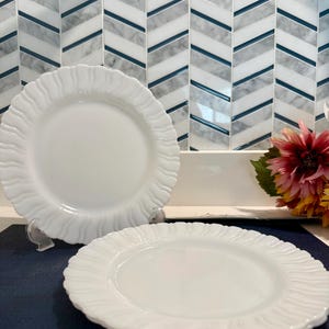 May include: Two white, ruffled-edge plates. The plates are on a dark blue surface, with a patterned tile backsplash in the background. The plates are round with a smooth center and a textured, wavy edge. A flower arrangement is visible in the background.