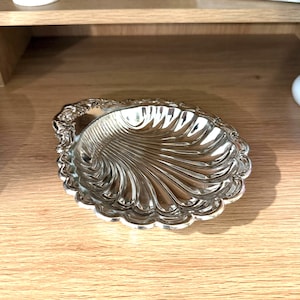May include: A polished silver-tone metal dish shaped like a scallop shell. The dish features a reflective, smooth interior with radiating fluted grooves and a decorative, ornate scalloped edge that adds texture to the overall design.