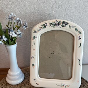 Puede incluir: Un marco de fotos de cerámica blanca con una parte superior arqueada, decorado con un diseño floral y de bayas en tonos verdes, rosas y morados. Un jarrón blanco con flores azules y blancas está a la izquierda.