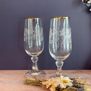 May include: Two clear champagne flutes with gold rims and etched grape vine designs. The glasses sit on a wooden surface with dried flowers and lavender. The background is a dark blue-purple.