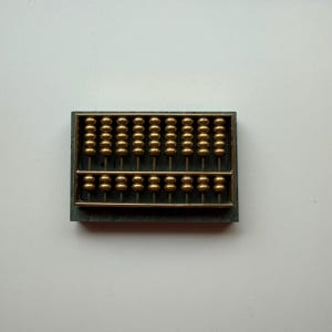 May include: A rectangular abacus featuring a dark gray frame and nine vertical metal rods. Each rod holds five gold-colored beads in the upper section and two gold-colored beads in the lower section, separated by a horizontal metal bar.